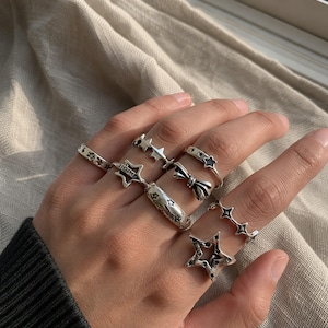 Può includere: Una collezione di anelli color argento indossati sulle dita, caratterizzati da vari design tra cui forme a stella, un fiocco e un anello con l'incisione HOPE. Gli anelli presentano una finitura metallica lucida con dettagli ossidati.