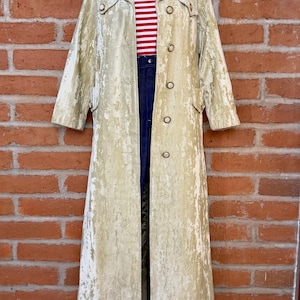 May include: A long, pale gold crushed velvet coat with a wide collar and a single row of round buttons. Underneath, a red and white horizontal striped shirt is paired with dark blue denim pants, all displayed on a mannequin form.