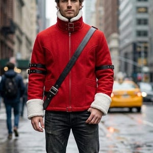 Könnte beinhalten: Eine leuchtend rote Jacke mit hohem Kragen und weißem Kunstfellfutter, weißen Kunstfellbündchen sowie schwarzen Lederriemen mit silbernen Schnallen an den Ärmeln und über der Brust. Die Jacke hat einen mittigen silbernen Reißverschluss.