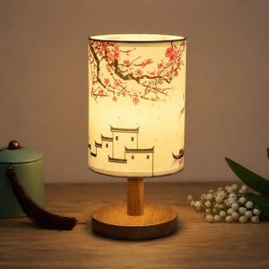 May include: A cylindrical table lamp with a light-coloured shade featuring a printed illustration of pink cherry blossoms and traditional buildings. The lamp has a circular wooden base and a central wooden stem, emitting a warm, soft glow.