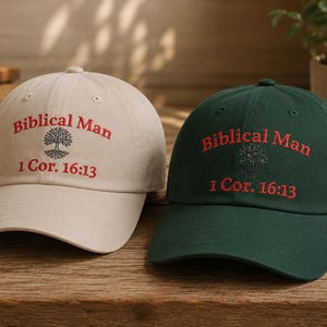 May include: Two baseball caps, one beige and one dark green, each featuring red embroidered text that reads Biblical Man and 1 Cor. 16:13, with a black embroidered tree graphic centered between the lines of text on the front of each cap.