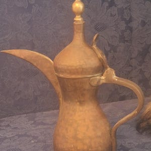 May include: A tall, hammered brass coffee pot featuring a long, slender spout, a curved handle, and a lid topped with a spherical finial. The metal surface has a muted, aged golden-brown finish with visible texture from the hand-hammering process.
