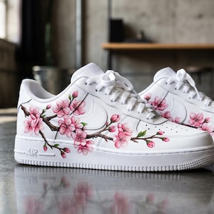 Könnte beinhalten: Ein Paar weiße Sneaker mit niedrigem Schaft und einem handgemalten Design aus rosa Kirschblüten auf braunen Zweigen, die sich über die Seiten erstrecken. Die Schuhe haben weiße Schnürsenkel, eine weiße Sohle und die Prägung AIR an der Ferse.
