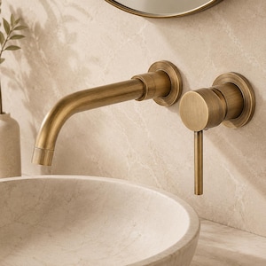 May include: A wall-mounted bathroom faucet with a brushed brass finish, featuring a curved spout and a separate cylindrical handle lever. The fixture is installed above a light-colored stone vessel sink against a matching marble wall.