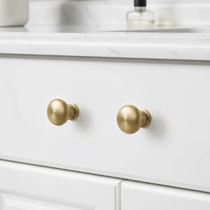May include: Two round cabinet knobs with a brushed brass finish attached to a white drawer front. The knobs feature a smooth, spherical shape with a subtle metallic sheen, providing a functional grip for opening the drawer in a kitchen or bathroom.