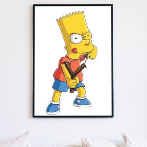 May include: A framed wall art print featuring a yellow cartoon character with spiky hair, wearing a red t-shirt and blue shorts, holding a wooden slingshot with a focused expression. The character has one eye squinted and tongue sticking out.