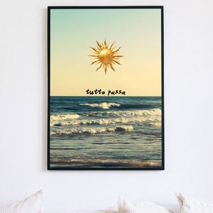 May include: A framed wall art print featuring a golden sun illustration centered above a blue ocean with white-capped waves. The text "tutto passa" is printed in black, lowercase letters between the sun and the horizon line of the sea.