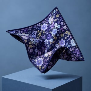 May include: A square pocket square featuring a dark navy blue background with a dense floral pattern in shades of light purple, lavender, and white. The fabric has a crisp, folded structure with a dark blue stitched border along the edges.
