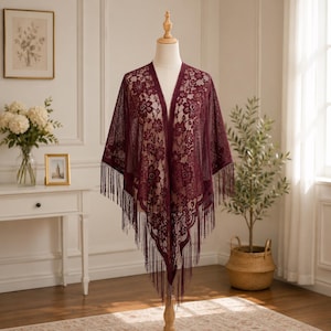 May include: A deep burgundy lace shawl featuring an intricate floral pattern throughout the fabric. The garment has an open front design and is finished with long, thin fringe detailing along the bottom edges, draped over a mannequin torso.