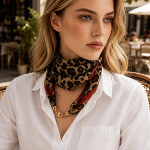 May include: A square scarf featuring a brown and black leopard print pattern with a dark red border, secured at the front with a gold-toned metal chain link connector, worn over a crisp white button-down shirt with a classic collar and chest pocket.