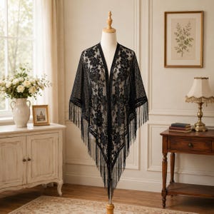 May include: A black lace shawl featuring an intricate floral pattern throughout the fabric. The garment has an open front design and is finished with long, thin fringe detailing along the bottom edges, creating a pointed, triangular silhouette.