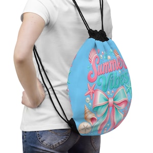 Peut inclure: Un sac à dos à cordon bleu clair avec le texte Summer Vibes en écriture rose et bleu sarcelle. Le motif comprend un grand nœud rose et bleu sarcelle, divers coquillages, une étoile de mer rose, une étoile de mer bleu sarcelle et des perles.