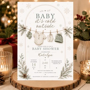 Könnte beinhalten: Eine weiße Babyparty-Einladungskarte mit dem Text Baby it's cold outside, einer Wäscheleine mit Body, Fäustlingen, Socken und Mütze, verziert mit Tannenzweigen, Schneeflocken und den Veranstaltungsdetails für Katelyn am 14. Dezember 2024.
