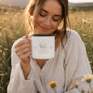 May include: A white enamel mug with a black rim featuring a floral illustration and the text "roaming slowly" and "wildflower bloom" printed in a rectangular frame on the front, with small floral accents scattered around the side of the mug.