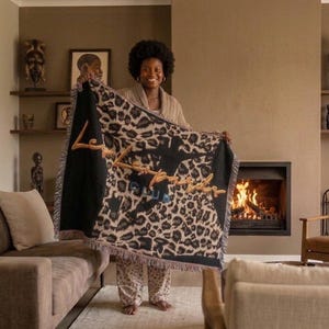 May include: A rectangular woven throw blanket featuring a black and tan leopard print pattern. The center displays a dark silhouette of a person and the embroidered text "Les Leopards CLUB" in gold and blue thread, with fringed edges on all sides.