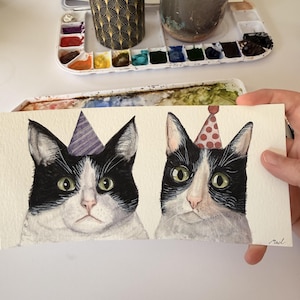 May include: A watercolor painting on paper featuring two black and white cats wearing party hats. The cat on the left wears a purple striped hat, while the cat on the right wears a red polka-dot hat. The artist's signature, Mad, is in the corner.