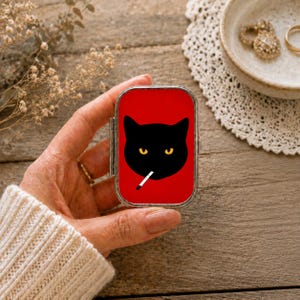 Pode incluir: Uma caixa de comprimidos retangular de metal com um fundo vermelho vibrante e uma ilustração gráfica de um rosto de gato preto. O gato tem olhos amarelos penetrantes e um cigarro branco na boca. O estojo tem uma moldura de metal prateado.