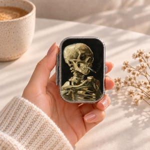 May include: A rectangular metal pill case featuring a reproduction of Vincent van Gogh's painting of a skeleton with a burning cigarette in its mouth. The case has a silver-toned metallic frame and a hinged lid with a glossy finish.