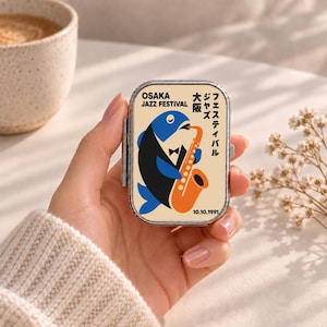 May include: A rectangular metal pill case with a cream-coloured lid featuring a graphic of a blue and black fish wearing a tuxedo and playing an orange saxophone. The text reads OSAKA JAZZ FESTIVAL, Japanese characters, and the date 10.10.1991.