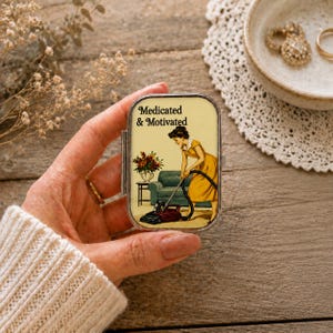 Op de afbeelding: Een rechthoekig metalen pillendoosje met een vintage-stijl illustratie van een persoon in een gele jurk die aan het stofzuigen is. De tekst Medicated & Motivated staat in het zwart boven de illustratie. Het doosje heeft een zilverkleurig metalen frame.