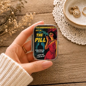 Könnte beinhalten: Ein rechteckiges Pillendöschen aus Metall mit einer Grafik im Vintage-Stil, die eine rauchende Frau in einem roten Kleid zeigt, bedruckt mit dem Text 'SHE'S ON... THE PILL', 'THE BLACK WIDOW WAITS FOR HER NEXT JOHN' und 'A SINFUL WEB OF LUST AND LIES'.