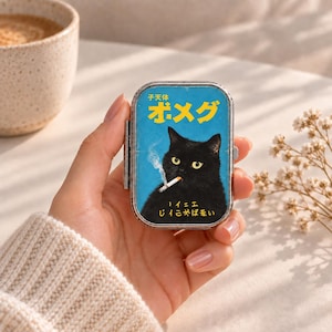 Pode incluir: Uma caixa metálica retangular com fundo azul e um gato preto segurando um cigarro aceso na boca. O design inclui texto japonês amarelo na parte superior e inferior, com uma borda metálica prateada fina ao redor de toda a caixa.