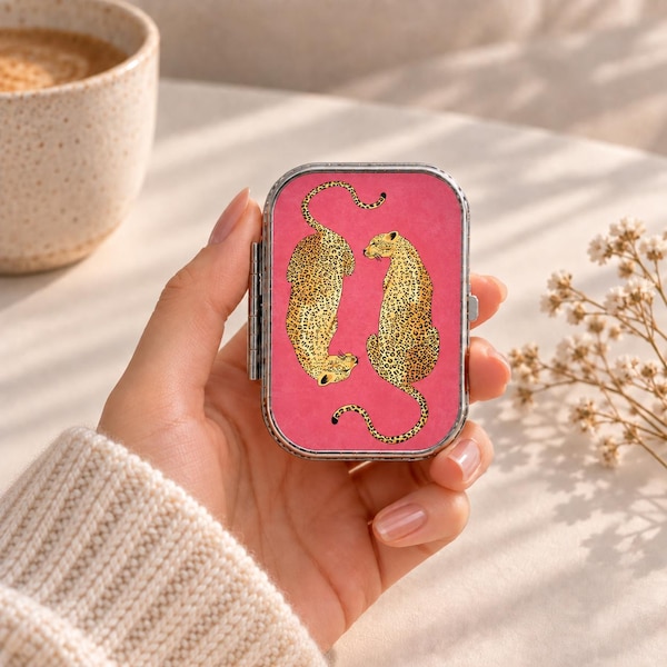May include: A rectangular metal compact case with a vibrant pink background featuring two illustrated leopards with yellow and black spotted fur. The leopards are positioned facing each other, with their long tails curving gracefully across the surface.