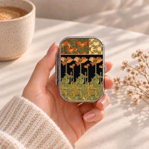 Puede incluir: Un pastillero rectangular de metal con un patrón floral decorativo de amapolas naranjas, tallos y hojas verdes sobre un fondo negro. La sección superior muestra un motivo floral más pequeño y un patrón geométrico de color crema.