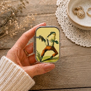 May include: A rectangular metal pill box featuring a colourful illustration of a green frog wearing a white shirt and brown trousers while holding a small firearm. The frog is depicted standing in a grassy field against a pale yellow background.