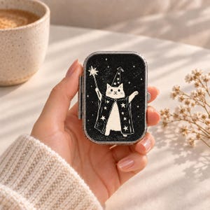 Könnte beinhalten: Eine rechteckige Pillendose aus Metall mit Klappdeckel, die auf schwarzem Grund eine weiße Illustration einer Katze mit Zaubererhut und Umhang zeigt, die einen sternförmigen Zauberstab hält. Die Katze ist von kleinen weißen Sternen umgeben.