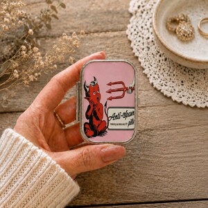 Puede incluir: Un pastillero rectangular de metal con tapa abatible que presenta un fondo rosa, un personaje de diablo rojo, un tridente rojo y el texto Anti-Spawn pills. La caja tiene un marco de metal plateado y un mecanismo de cierre seguro.
