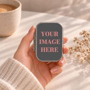 Peut inclure: Un boîtier compact rectangulaire en métal argenté avec un couvercle à charnière. La surface avant est gris foncé et présente le texte rose YOUR IMAGE HERE centré au milieu. Le boîtier a un cadre métallique et une charnière visible à gauche.