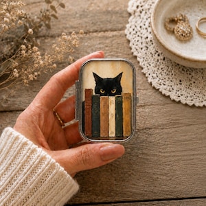 Pode incluir: Uma caixa metálica retangular com dobradiça que apresenta uma ilustração impressa de um gato preto com olhos amarelos espreitando sobre uma fila de cinco livros antigos. A caixa tem uma moldura prateada e uma tampa transparente.
