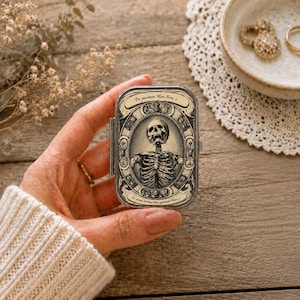 Könnte beinhalten: Eine rechteckige Metalldose mit einer Illustration im Vintage-Stil, die einen menschlichen Skelettorso in einem verzierten ovalen Rahmen zeigt. Das Design umfasst dekorative Schnörkel und den lateinischen Text Per peccatum Mors Rom 5.