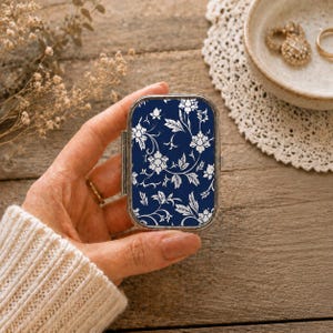 Puede incluir: Un pastillero rectangular de metal con fondo azul marino oscuro y un patrón repetitivo de flores y enredaderas blancas. El estuche tiene un marco de metal plateado y una tapa con bisagras que muestra un diseño botánico detallado.