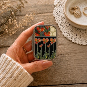 Könnte beinhalten: Eine rechteckige Pillendose aus Metall mit einem dekorativen Design aus orangefarbenen Mohnblumen auf schwarzem Grund. Der obere Bereich zeigt ein Muster aus roten Mohnblumen und einem cremefarbenen Gitter. Das Gehäuse hat einen silberfarbenen Rahmen.