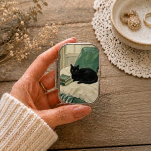 Könnte beinhalten: Eine rechteckige Pillendose aus Metall mit einer gemalten Illustration einer schwarzen Katze, die auf einer weißen Oberfläche neben zwei kleinen Büchern liegt, wobei im Hintergrund ein grüner Stuhl zu sehen ist. Der Deckel ist klappbar und silberfarben.