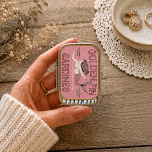 Peut inclure: Une boîte métallique rectangulaire avec une étiquette rose portant le texte GOLDEN FIN SARDINES. L'étiquette présente un dessin au trait noir d'une sirène et une rangée de petits poissons en bas. La boîte est tenue par une main en manche tricotée.