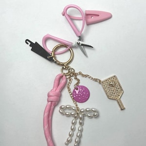 May include: A keychain featuring a gold-tone ring attached to a pink paracord loop, a pink miniature scissor charm, a small purple circular charm, a gold-tone pickleball paddle charm, and a bow made of small white faux pearls.