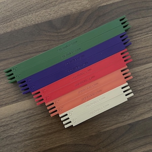 May include: A set of ten flat, rectangular plastic bars in green, purple, red, orange, and cream. Each bar features notched ends and is printed with its length in inches, ranging from 5" to 9", and the website text srqstudios.com.