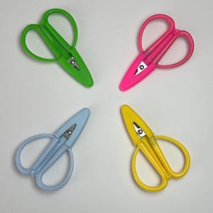 May include: Four pairs of thread snips with plastic handles and protective caps, arranged in a square formation. The colors include bright green, vibrant pink, light blue, and yellow. Each tool features a metal blade tip and a spring-loaded mechanism.