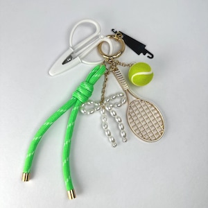 May include: A keychain featuring a gold-tone metal tennis racket charm, a small yellow-green tennis ball, a white faux pearl bow, and a bright neon green braided cord with gold-tone metal tips, all attached to a white plastic scissor-shaped clip.