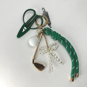 Puede incluir: Un llavero con un dije de palo de golf dorado, una pelota de golf blanca texturizada, un pequeño lazo de perlas de imitación y unas tijeras de hilo verdes. Incluye un cordón trenzado verde con motas blancas y terminales metálicos dorados.