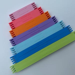 May include: A set of ten rectangular plastic weaving loom bars in a gradient of colors including pink, orange, purple, blue, and green. Each bar features notched ends and is printed with its length in inches from 4 to 10 and the text srqstudios.com.