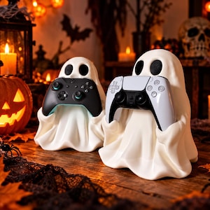 Könnte beinhalten: Zwei weiße geisterförmige Controller-Ständer aus glattem Material. Der linke Geist hält einen schwarzen kabellosen Game-Controller mit türkisfarbenen Akzenten, während der rechte Geist einen weiß-schwarzen kabellosen Controller hält.