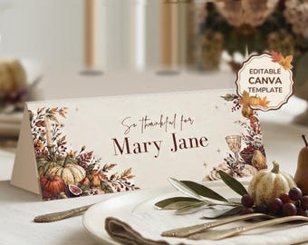 Può includere: Un segnaposto in carta piegata con sfondo color crema, decorato con illustrazioni autunnali di zucche, foglie e frutta. Il testo recita "So thankful for Mary Jane" in un carattere marrone scuro corsivo e serif. Un badge riporta "EDITABLE CANVA TEMPLATE".