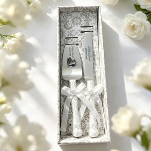 Op de afbeelding: Een zilveren taartmes en taartschep in een doos met patroon. De handvatten zijn omwikkeld met wit kant en satijnen lint met een klein hartje. De metalen bladen zijn gegraveerd met de tekst Lily & Jack, June 5th, 2026 en een afbeelding van een paar.