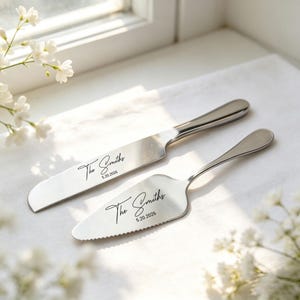 May include: A silver-toned cake knife and server set with a polished finish. Both pieces are engraved with the text The Smiths and the date 5.20.2026 in a black script font. The server features a serrated edge on one side for cutting.