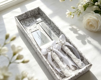 Könnte beinhalten: Ein Tortenmesser- und Tortenheber-Set aus Silber in einer gemusterten Schachtel. Beide Teile haben weiße florale Griffe, weiße Satinbänder und herzförmige Anhänger. Die Klingen sind mit dem Text Alexandra & Jace und dem Datum 24.07.27 graviert.