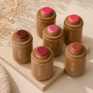 May include: Six cylindrical cosmetic containers in a tan shade, each featuring the brand name rhode printed vertically in white. The containers have circular, raised tops in varying shades of pink, red, and berry, arranged in a cluster.